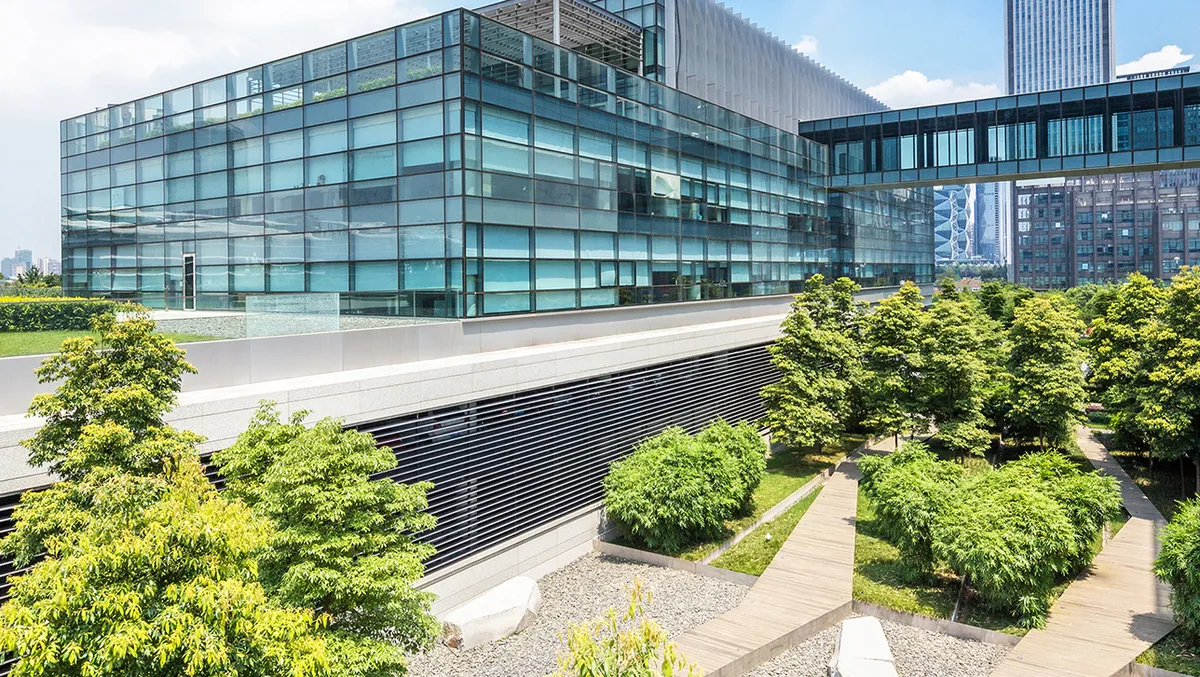 Modernes Bürogebäude mit Glasfassade und begrüntem Außenbereich. Vor dem Gebäude befinden sich gepflegte Bäume, Sträucher und ein Holzsteg, der sich durch die Anlage schlängelt. Eine gläserne Brücke verbindet die Gebäudeteile, im Hintergrund sind weitere Hochhäuser der Stadt zu sehen.