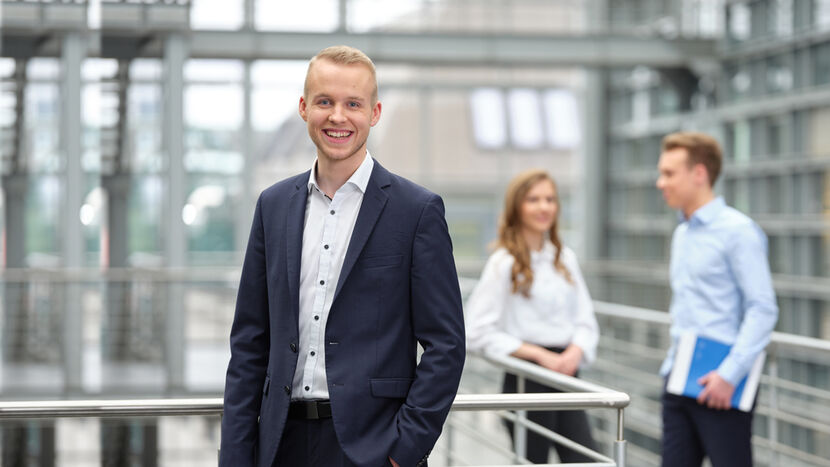 Ein junger Mann in Businesskleidung steht im Innenhof eines Bürogebäudes und blickt lächeln aus dem Bild, im Hintergrund zwei junge Menschen im Gespräch