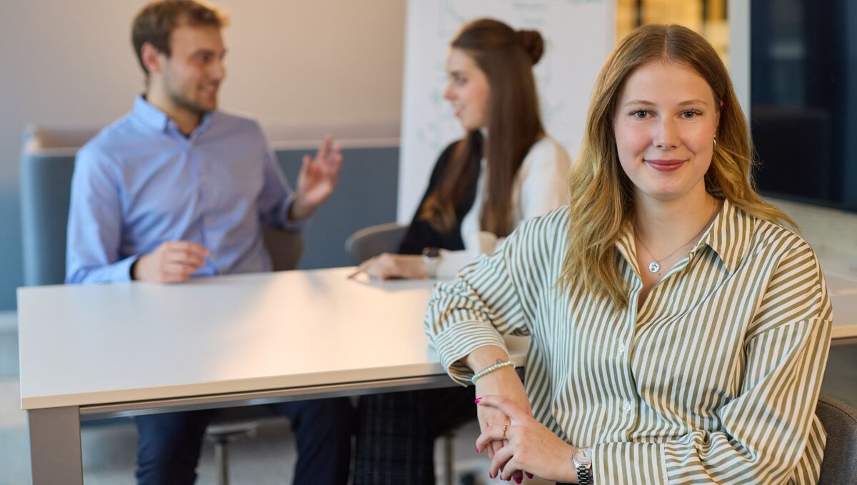 Werkstudierende bei der DZ HYP Drei Werkstudierende. Zwei von ihnen befinden sich im Hintergrund und unterhalten sich. Eine junge Frau sitzt vorne und blickt freundlich in die Kamera.