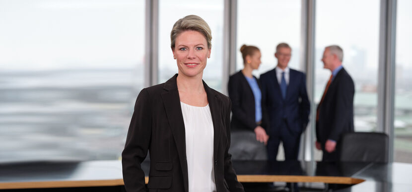 Eine stehende Frau in Businesskleidung blickt freundlich lächelnd aus dem Bild, im Hintergrund stehen drei Menschen in Businesskleidung im Gespräch miteinander in einem Konferenzraum vor einer Fensterfront