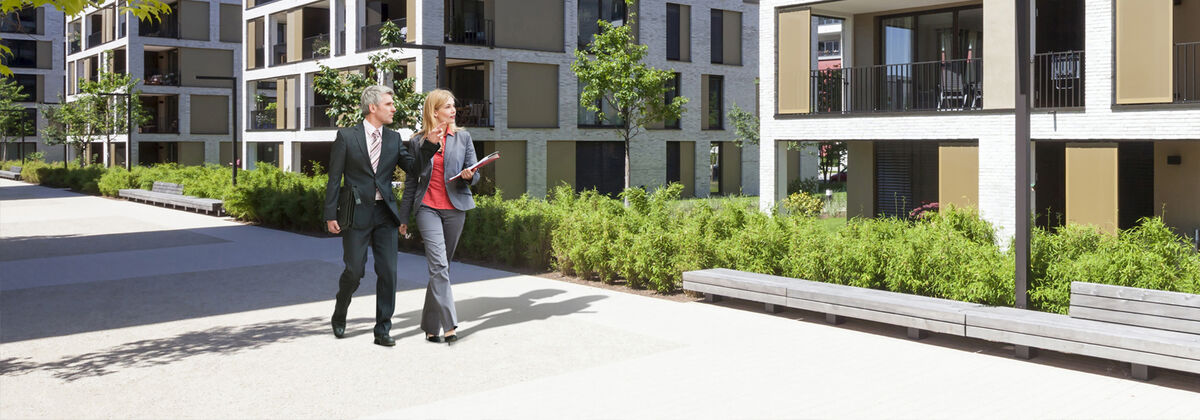 A man and a woman in business attire walk past newly built apartments while talking. The man points to a building.