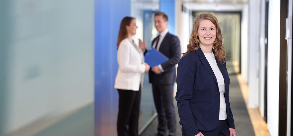 Eine stehende Frau in Businesskleidung blickt freundlich lächelnd aus dem Bild, im Hintergrund stehen drei Menschen in Businesskleidung im Gespräch miteinander in einem Büroflur