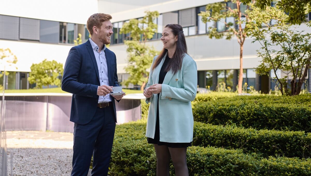 Ein Mann und eine Frau stehen in einem grünen Innenhof und unterhalten sich. 