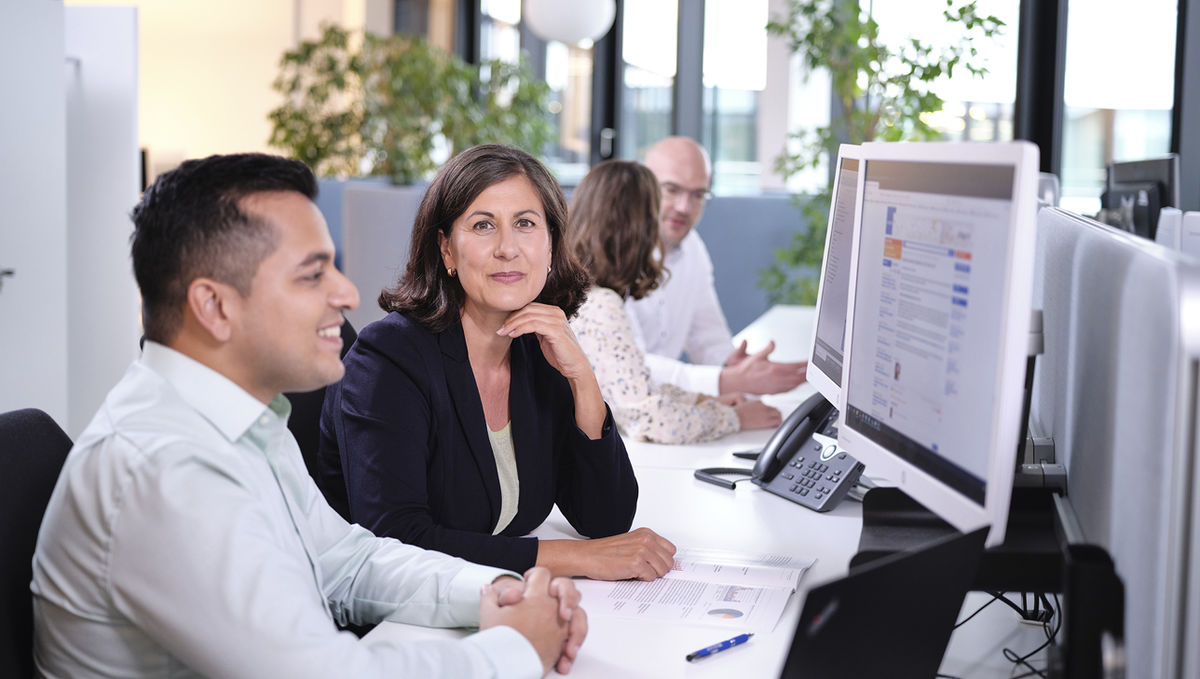 Eine sitzende Frau in Businesskleidung blickt freundlich lächelnd aus dem Bild, im Hintergrund sitzen zwei Menschen in Businesskleidung im Gespräch miteinander an einem Bürotisch