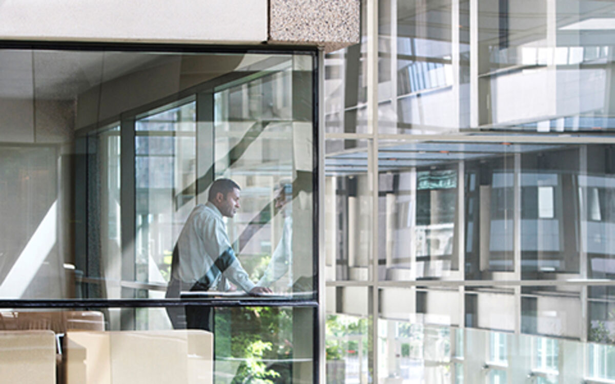 A man in a business shirt and trousers looks out of a glassed-in room into the courtyard of a glass office building
