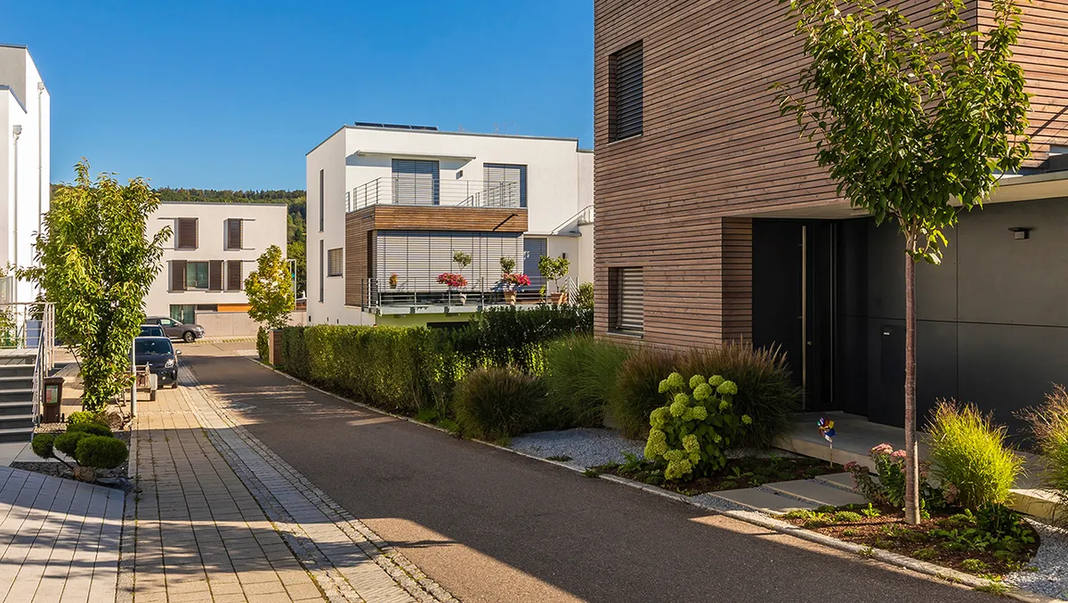 Ruhige Wohnstraße mit modernen Einfamilienhäusern in kubischer Architektur. Die Gebäude haben helle Fassaden, teils mit Holzverkleidung und großen Fenstern. Begrünte Vorgärten, Bäume und Pflanzbeete verleihen dem Viertel eine gepflegte und naturnahe Atmosphäre bei sonnigem Wetter.