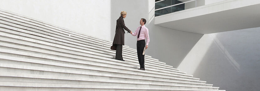 Eine Frau und ein Mann in Businesskleidung auf der großen, hellen Treppe eines Bürogebäudes, lächelnd und Hände schüttelnd