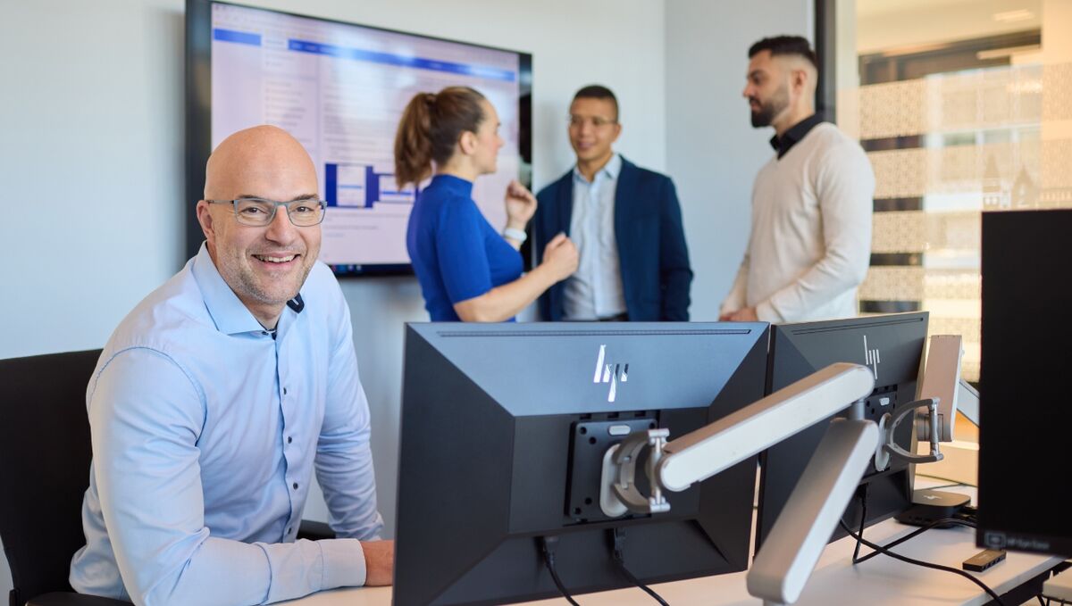 Karriere in der IT Ein Mann sitzt im Vordergrund und lächelt in die Kamera, im Hintergrund stehen eine Frau und zwei weitere Männer.