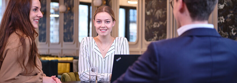 Eine junge Frau in schwarz-weiß gestreifter Bluse blickt lächelnd aus dem Bild, vor ihr steht ein aufgeklappter Laptop, rechts von ihr sitzt eine junge Frau, ihr gegenüber ein junger Mann