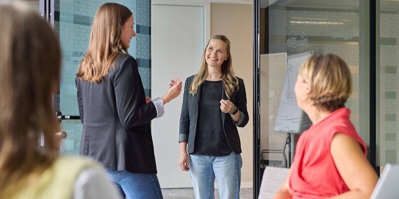 Vier Frauen stehen in einem Konferenzraum und unterhalten sich. 