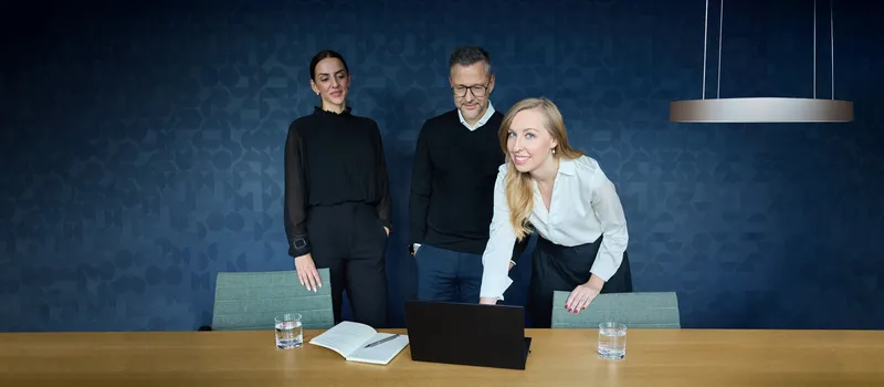 Zwei Frauen und ein Mann stehen in einem Konferenzraum, die vordere Frau guckt in die Kamera.