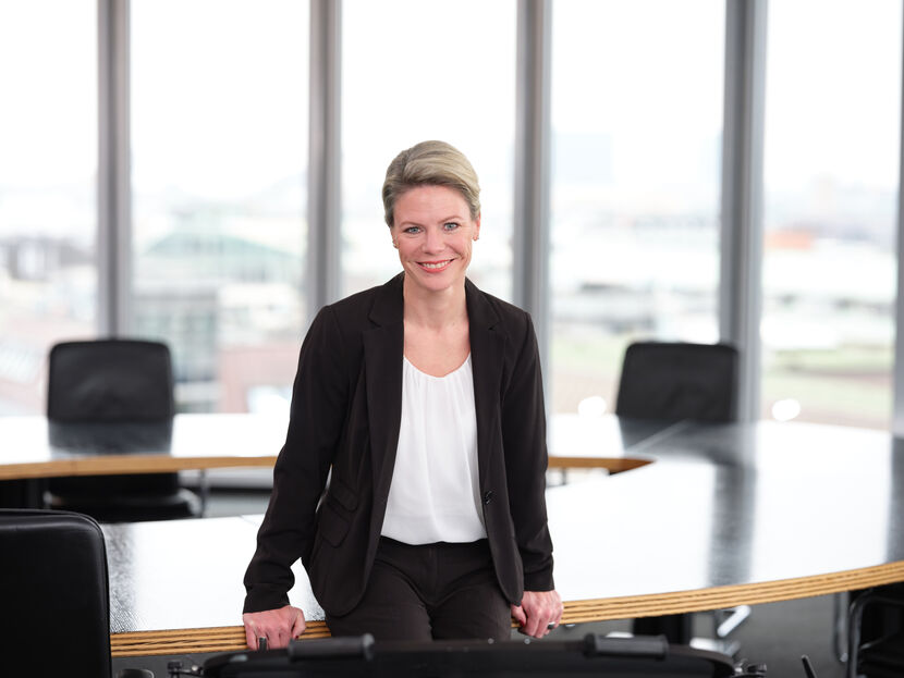 Eine Frau in Businesskleidung sitzt locker auf einem Konferenztisch und blickt freundlich lächelnd aus dem Bild, im Hintergrund eine Fensterfront