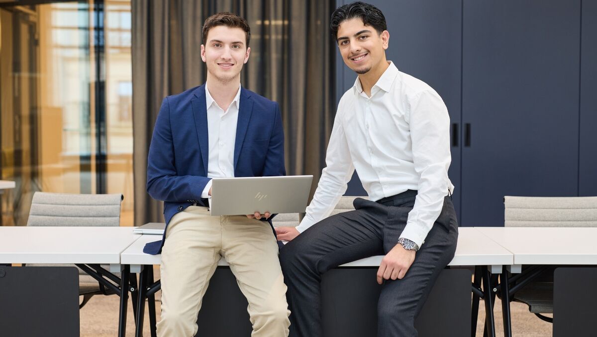 Zwei Nachwuchskräfte lehnen an einem Tisch und lächeln freundlich in die Kamera