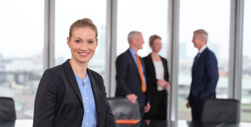 Eine Frau in Businesskleidung blickt freundlich lächelnd aus dem Bild, im Hintergrund stehen drei Menschen in Businesskleidung im Gespräch miteinander in einem Konferenzraum vor einer Fensterfront