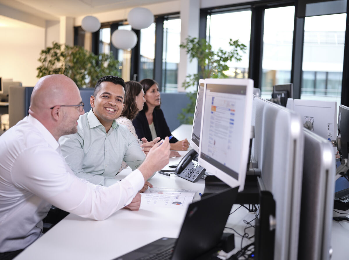 Vier Mitarbeiterinnen und Mitarbeiter sitzen in einer Reihe vor PC-Bildschirmen, im Hintergrund eine Fensterfront. Ein Mann blickt lächelnd aus dem Bild