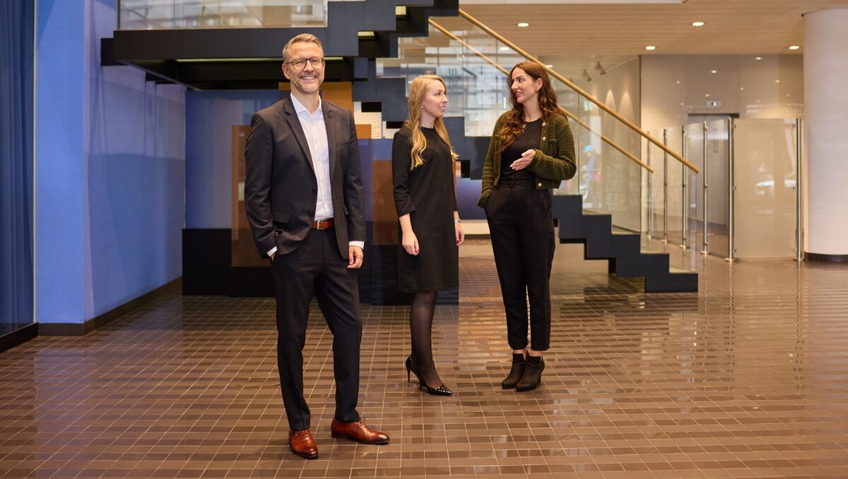 Professional bei der DZ HYP Ein Mann steht im Vordergrund, zwei Frauen im Hintergrund. Alle Personen befinden sich vor einer Treppe in einem Foyer von einem Bürogebäude.