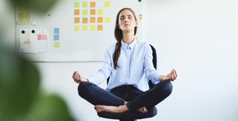 Eine junge Frau in Businesskleidung sitzt in Meditationshaltung auf einem Bürostuhl, im Hintergrund eine weiße Wand, daran eine Pinnwand mit bunten Post-its