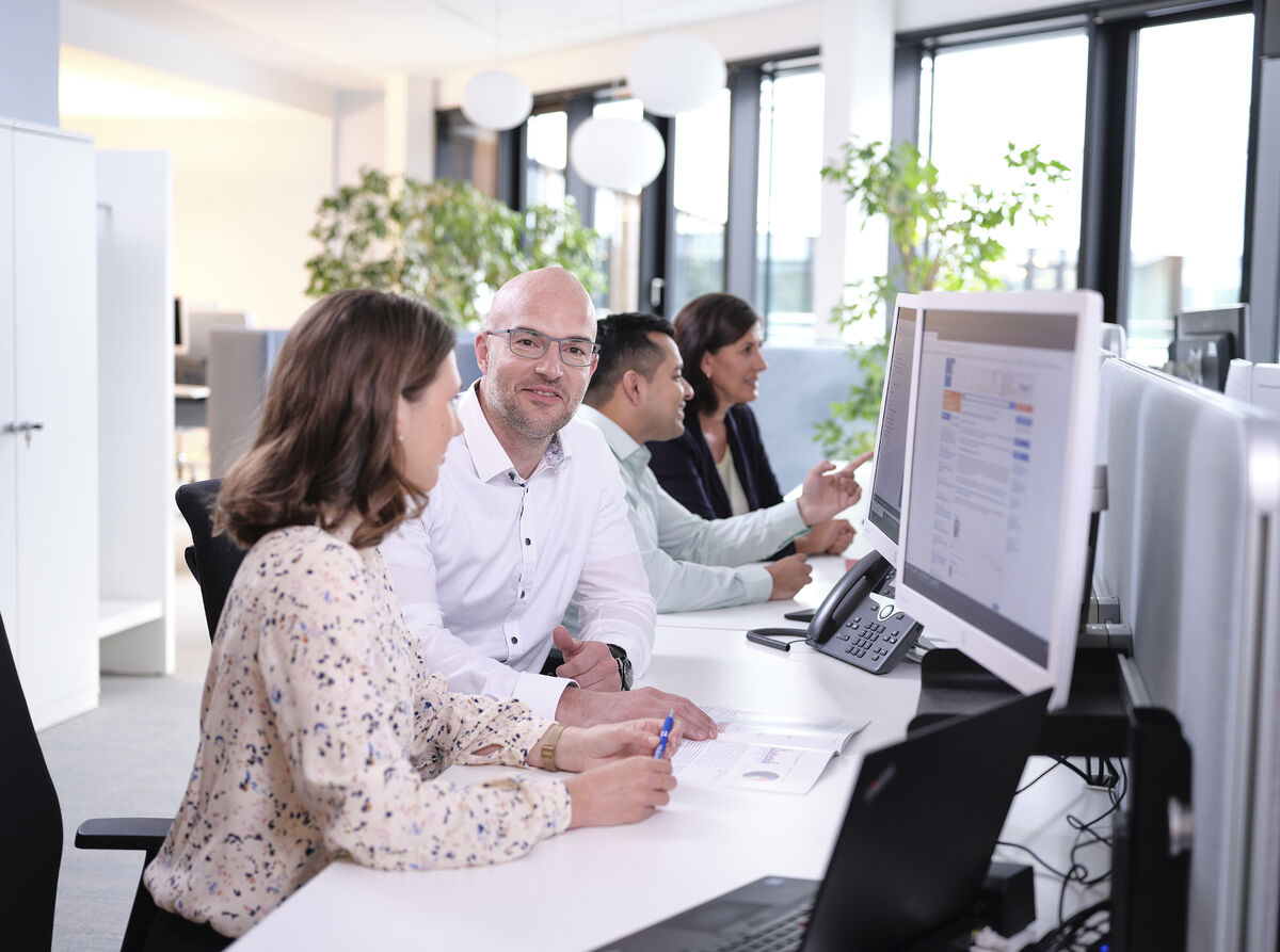 Vier Mitarbeiterinnen und Mitarbeiter sitzen in einer Reihe vor PC-Bildschirmen, im Hintergrund eine Fensterfront. Ein Mann blickt freundlich aus dem Bild