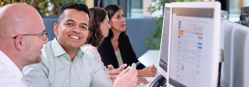 Vier Mitarbeiterinnen und Mitarbeiter sitzen in einer Reihe vor PC-Bildschirmen. Ein Mann blickt lächelnd aus dem Bild