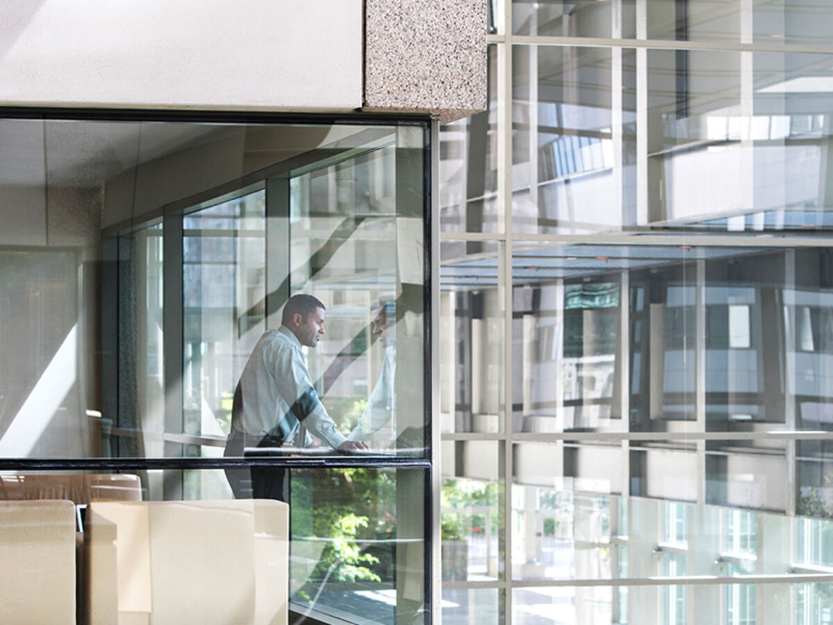 Ein Mann in Businesshemd und -hose blickt aus einem verglasten Raum in den Innenhof eines gläsernen Bürogebäudes