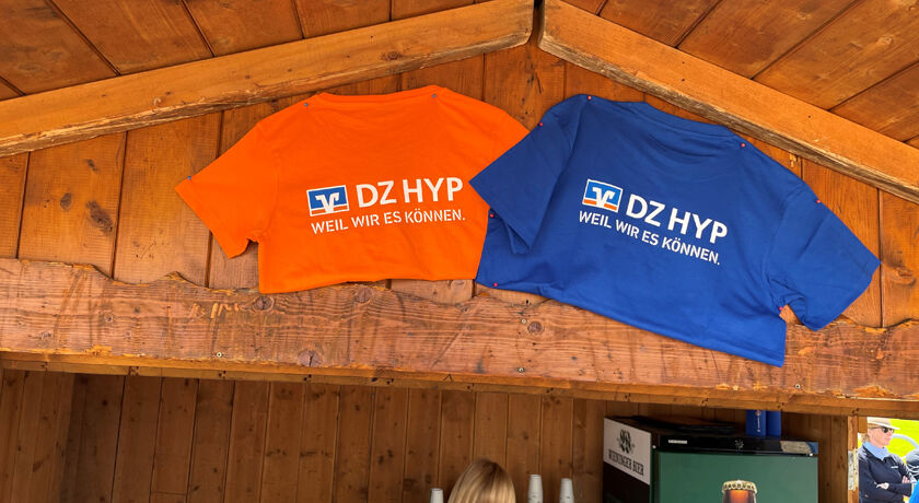 Ein orangefarbenes und blaues DZ HYP T-Shirt hängt festgenagelt an einer Holzhütte. 