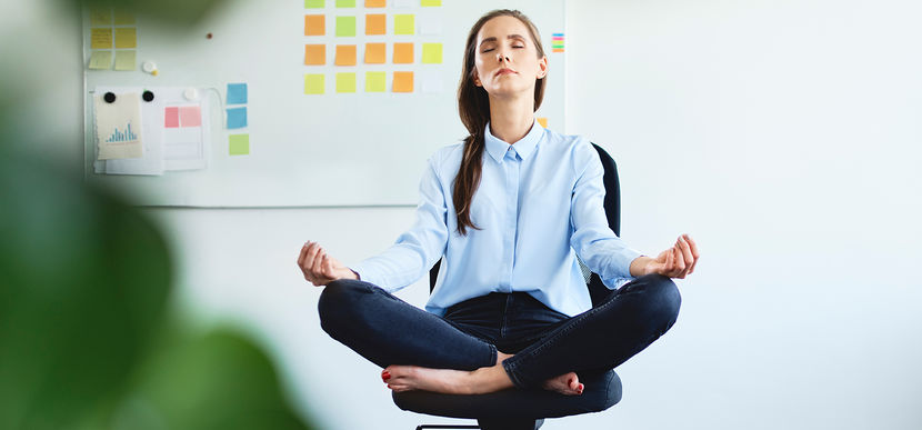 Eine junge Frau in Businesskleidung sitzt in Meditationshaltung auf einem Bürostuhl, im Hintergrund eine weiße Wand, daran eine Pinnwand mit bunten Post-its