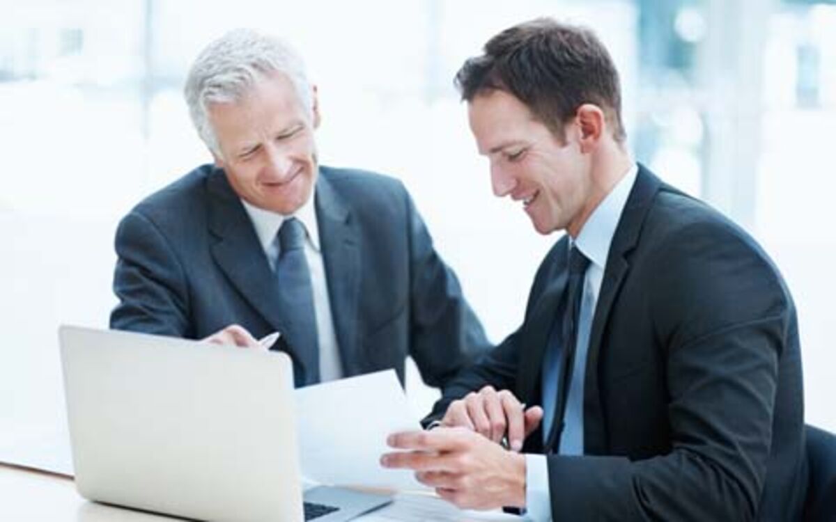 Two men sit smiling next to each other at a table in front of an open laptop. The older one points to the paper that the younger one is holding in his hand.