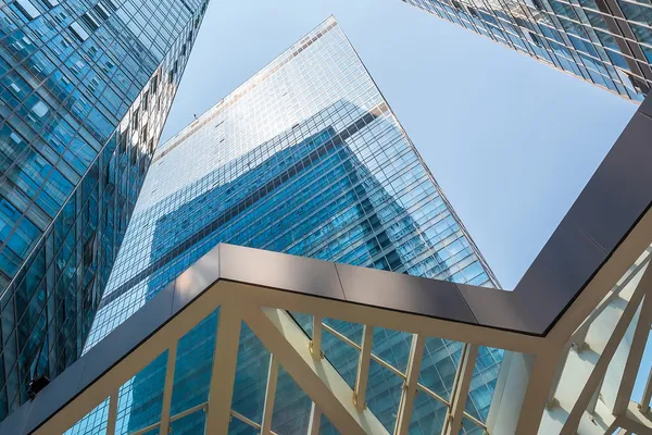 Blick nach oben zwischen modernen Glashochhäusern mit blauem Himmel und gläserner Fußgängerbrücke im Vordergrund