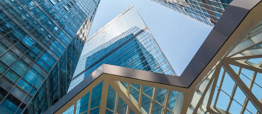 View upwards between modern glass skyscrapers with blue sky and glass pedestrian bridge in the foreground