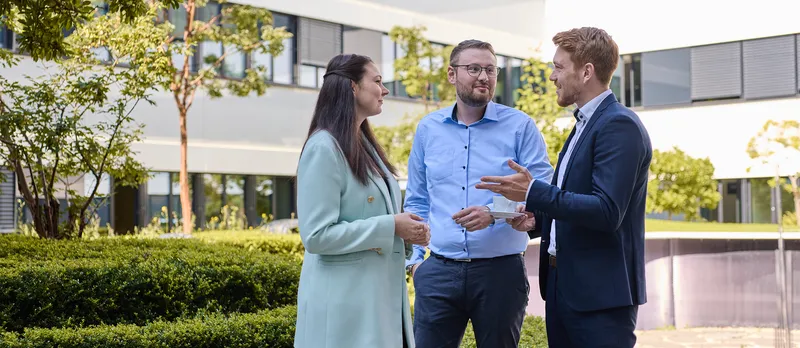 Eine Frau und zwei Männer stehen im grünen Innenhof und unterhalten sich. 