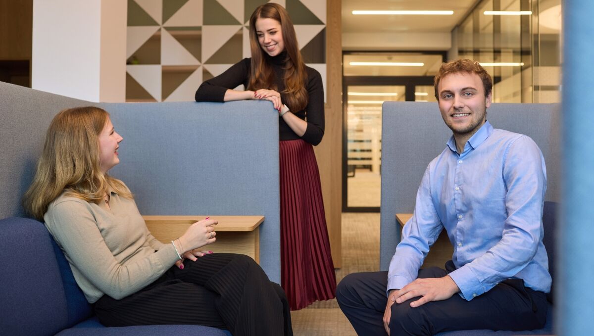Werkstudierende bei der DZ HYP Drei Werkstudierende sitzen in einer Lounge Ecke auf der Bürofläche. Zwei junge Frauen unterhalten sich im Hintergrund. Ein junger Mann blickt freundlich in die Kamera.