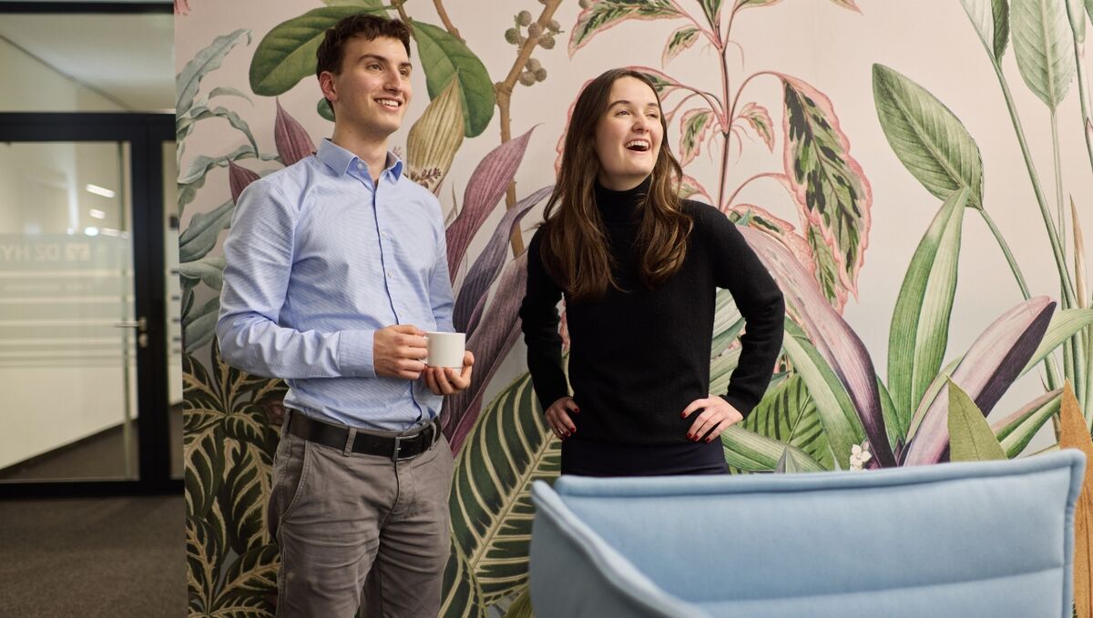 Zwei Auszubildende stehen vor einer farbigen Wand, die mit Blumen verziert ist und lachen freundlich